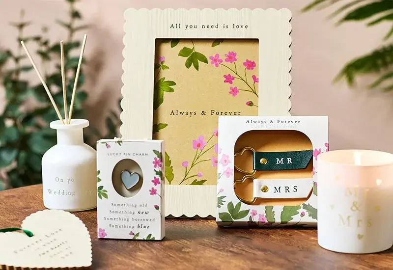 Wedding-themed items on a wooden surface, including a reed diffuser, picture frame with 'All you need is love' and 'Always & Forever', a 'Lucky Pin Charm' box, 'MR' and 'MRS' keychains, and a 'Mr & Mrs' candle holder, with green foliage in the background.